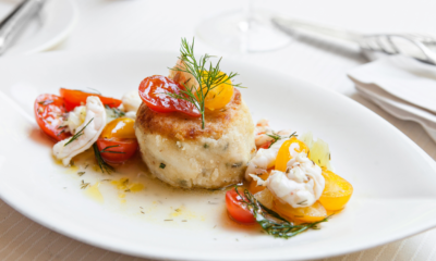 A mouthwatering crab cake plated with lemon remoulade, fresh greens, and a summer cocktail — one of many worth traveling for this year.
