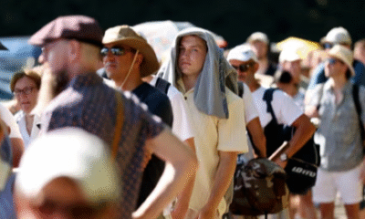 A group of people in england stand in a long queue under intense sunlight; many are wearing hats or using shirts and umbrellas for shade, indicating extreme heat conditions. A group of people stand in a long queue under intense sunlight; many are wearing hats or using shirts and umbrellas for shade, indicating extreme heat conditions