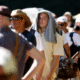 A group of people in england stand in a long queue under intense sunlight; many are wearing hats or using shirts and umbrellas for shade, indicating extreme heat conditions. A group of people stand in a long queue under intense sunlight; many are wearing hats or using shirts and umbrellas for shade, indicating extreme heat conditions