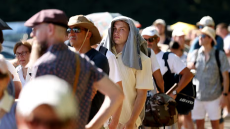 A group of people in england stand in a long queue under intense sunlight; many are wearing hats or using shirts and umbrellas for shade, indicating extreme heat conditions. A group of people stand in a long queue under intense sunlight; many are wearing hats or using shirts and umbrellas for shade, indicating extreme heat conditions