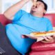 Overweight man eats pizza while sitting on couch at home
